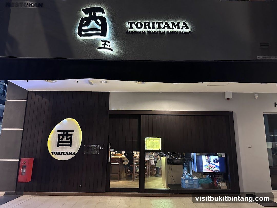 The exterior of a Japanese restaurant called Toritama. The storefront features dark wooden paneling and large glass doors. A prominent oval sign with the restaurant's logo and name is displayed on the left side of the entrance. Above the entrance, the restaurant's name "TORITAMA" is illuminated in white, with the words "Japanese Yakitori Restaurant" below it. A red fire extinguisher is mounted on the wall to the left of the entrance. Through the glass doors, a glimpse of the interior seating and a display screen showing food can be seen.