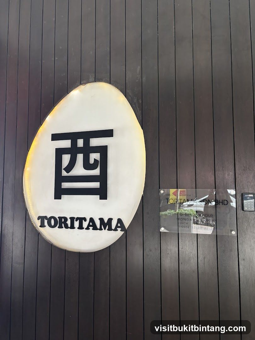 A white oval sign with a black Japanese character and the word "TORITAMA" in black letters is mounted on a dark wooden wall. To the right of the sign is a clear acrylic plaque displaying the business name "TORITAMA SDN BHD" and operating hours.