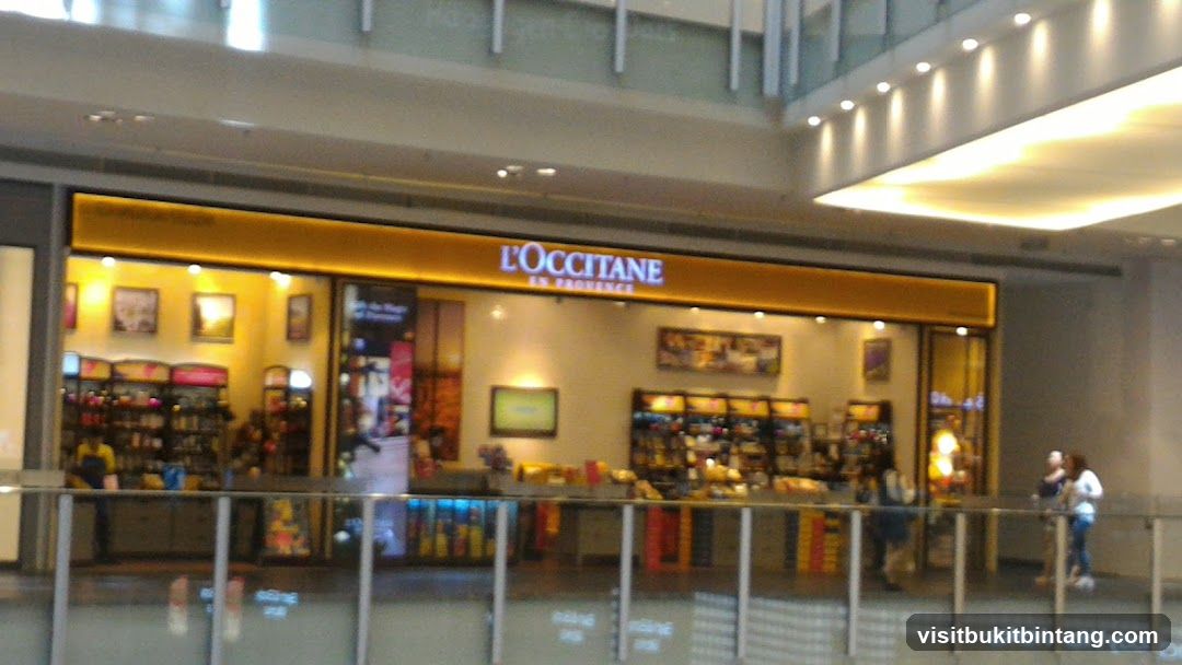 The storefront of L'OCCITANE en Provence is visible, featuring a bright orange awning with the store's name illuminated in white. The interior of the store displays shelves stocked with various products, and there are framed pictures on the walls. Several customers are seen browsing inside and near the entrance.