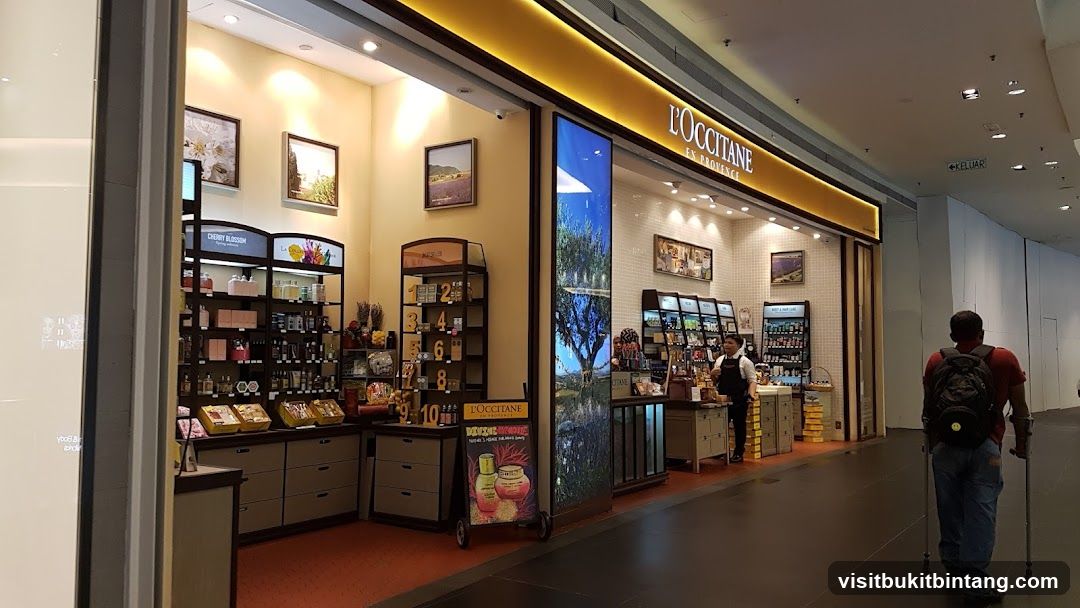 The exterior of a L'Occitane en Provence store in a mall. The store features a bright yellow illuminated sign with the brand name. Inside, shelves are stocked with various beauty and skincare products. A digital display shows a scenic landscape. A store employee is visible behind the counter. A man with crutches walks past the storefront.