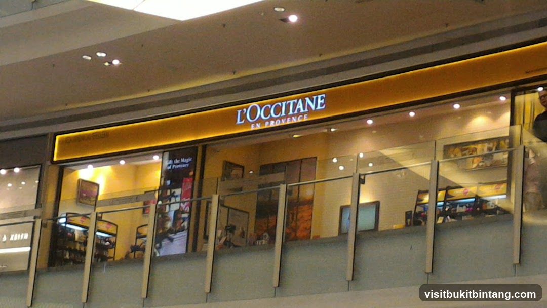 The exterior of a L'Occitane en Provence store is visible, with its prominent orange signage displaying the brand name in white lettering. The store appears to be located within a mall or shopping center, as indicated by the overhead lighting and the multi-level structure. Through the glass facade, shelves stocked with various products are visible, along with some decorative elements and displays. A person can be seen standing inside the store on the right side of the image.