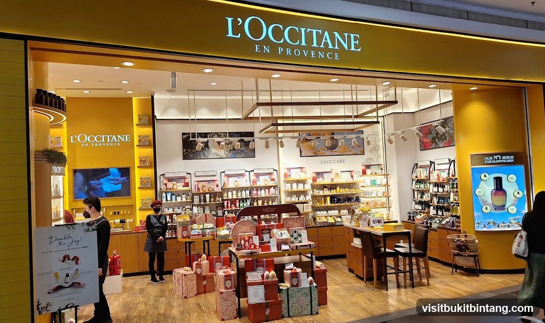 The exterior of a L'Occitane En Provence store, with its signature yellow branding and prominent signage. The store is filled with various skincare and beauty products displayed on shelves and tables. Two employees are visible inside the store, one near the entrance and another behind a counter. A promotional display for "Our N°1 Serum for Glowing Skin" is featured on the right side of the storefront.