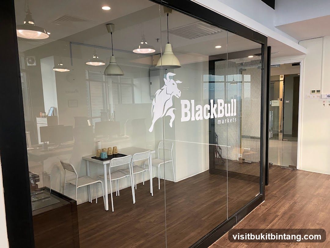 A view of the interior of an office space through a glass door and wall. The logo for BlackBull Markets is visible on the glass. Inside, there are desks with computers, a small table with chairs, and lighting fixtures.