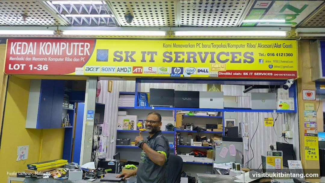 A computer shop with a yellow and red sign that says "KEDAI KOMPUTER" and "SK IT SERVICES". The sign also lists brands like Acer, Sony, AMD, ATI, Compaq, Nvidia, HP, Intel, and Dell. Below the sign, there are shelves with laptops and computer parts. A man is giving a thumbs up in front of the shelves. The shop is located in Kuala Lumpur, Malaysia.