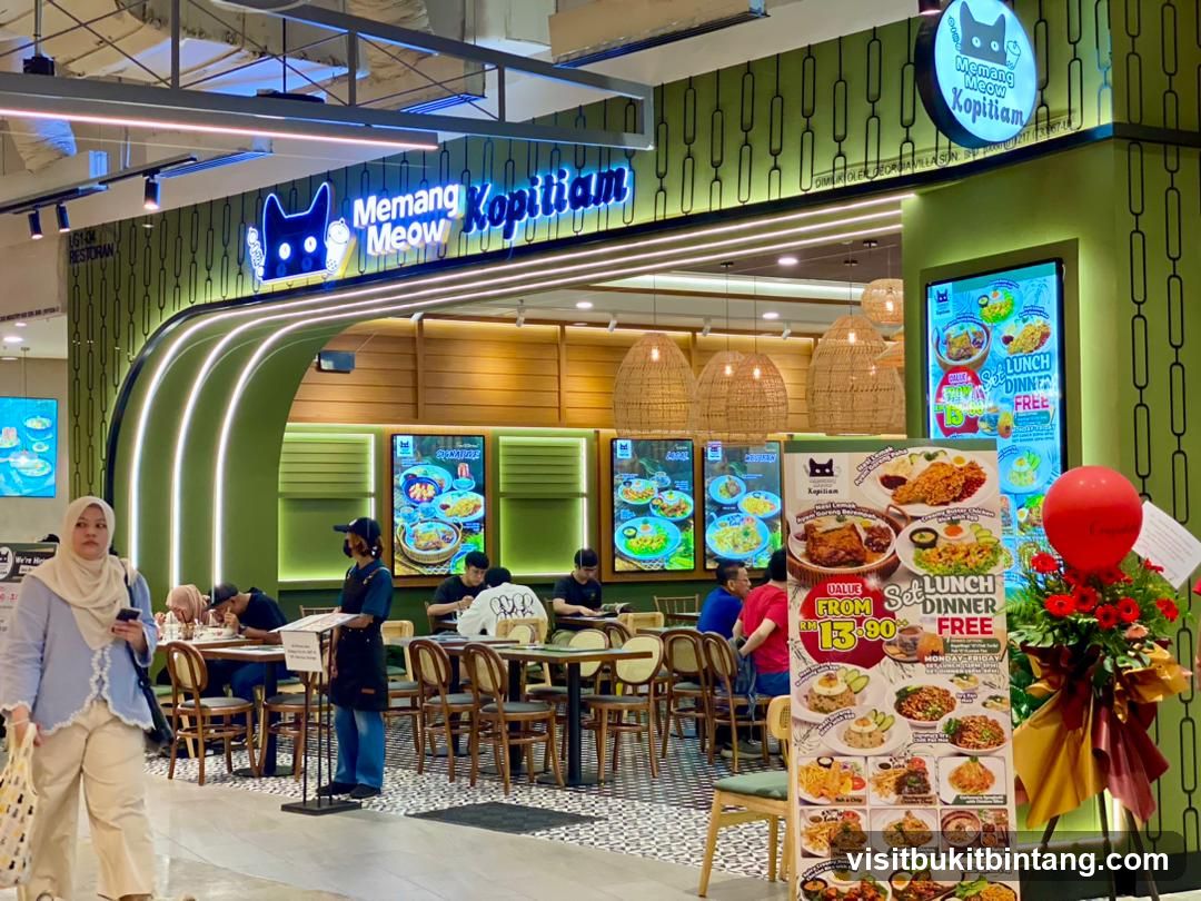 The exterior of a brightly lit restaurant with a green and wood-paneled facade. The restaurant's name, "Memang Meow Kopitiam," is displayed prominently in blue neon lettering above the entrance, accompanied by a cartoon cat logo. Inside, tables and chairs are visible, with patrons seated and dining. A large promotional poster featuring various dishes and meal deals is displayed to the right of the entrance.