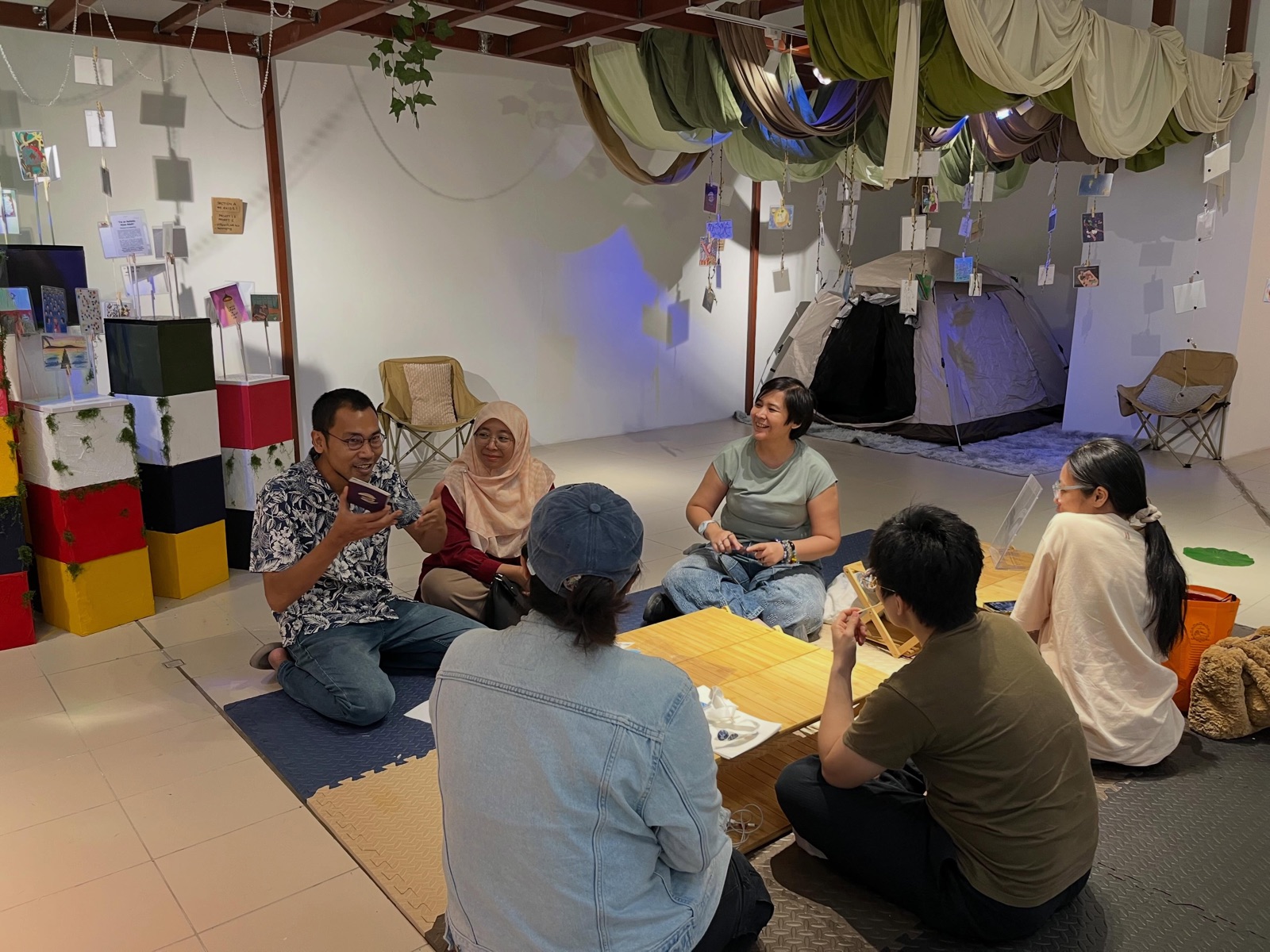 Pengunjung duduk berbual atas tikar di The Ways We Human, GMBB Aras 3, khemah dan kain tergantung di belakang